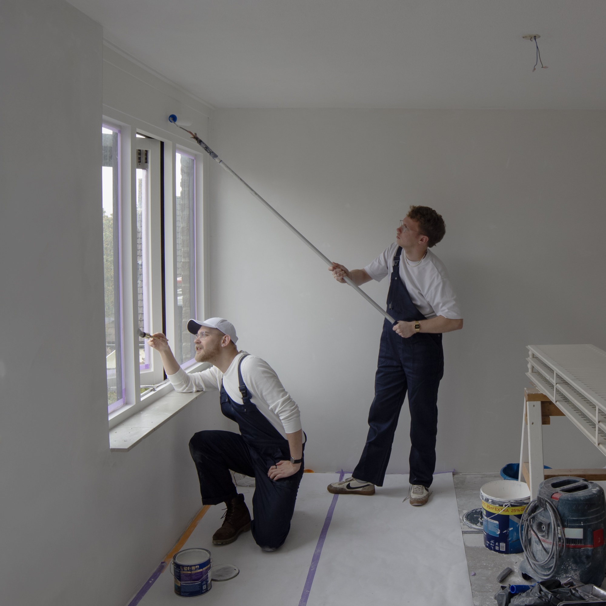 Twee Wijzen in tuinbroeken, druk aan het schilderen in een lege kamer.